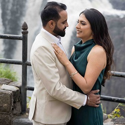 A couple posing closely outdoors by a stone railing and waterfall, with the woman in a green dress and the man in a beige suit, during a pre-wedding photoshoot.