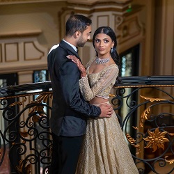 A groom in a black suit and a bride in a gold embellished gown pose together at an ornate indoor wedding venue railing.