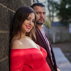 A smiling woman in a red off-shoulder dress posing outdoors beside a man in a suit, with a softly blurred background.