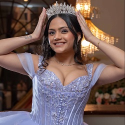Smiling young woman wearing a lavender Sweet 16 gown and tiara, posing indoors with her hands on her head.